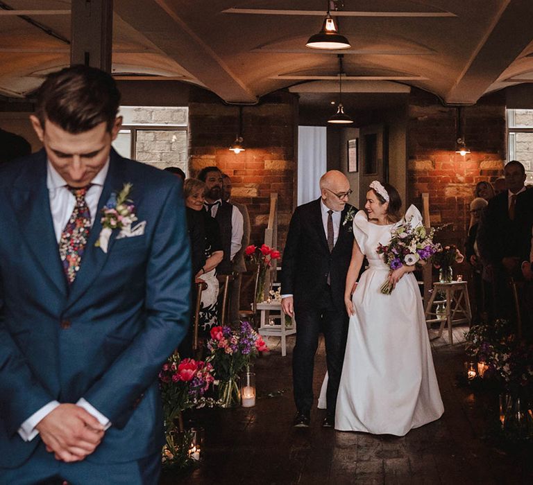 Father of the bride in black suit and burgundy tie walks the bride down the aisle at Bowers Mill with colourful spring flowers as the groom waits