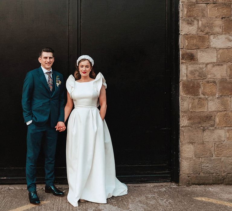 Bride in Jesus Peiro wedding dress with statement sleeves and pockets and white headband accessory holding hands with groom in navy blue suit and floral tie