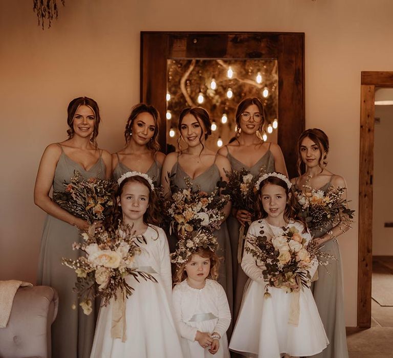 Bridesmaids holding autumnal coloured bouquets with flower girls in white dresses with cardigans and headbands or flower crowns