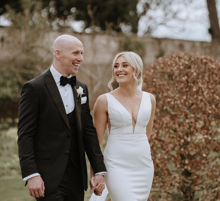 Bride and groom walk holding hands with groom in black tie and bride in fitted deep v button back wedding dress from Made With Love