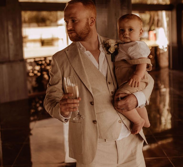 Groom in relaxed open shirt carries baby who wears white shirt and nude dungarees 