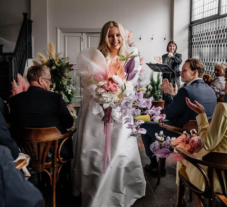 Bride in a JESUS PEIRO wedding dress with long sheer sleeves descending down the aisle holding a pastel pink, lilac and white wedding bouquet 