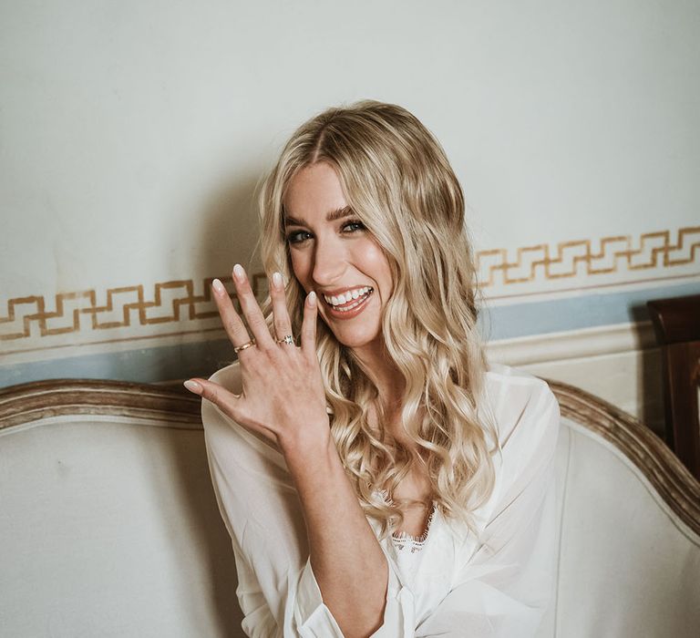 Bride wears white pyjamas and dressing gown with lace trim and her blonde hair in loose curls on the morning of her wedding day