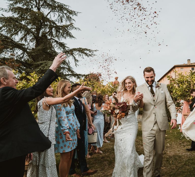 Bride holding colourful bouquet & groom walk through confetti after Tuscan wedding ceremony at the Villa Catignano 