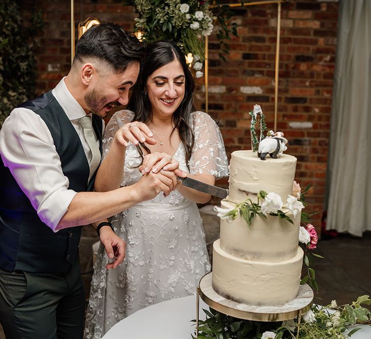 Bride & groom cut three tier naked wedding cake finished with white frosting and floral decor 