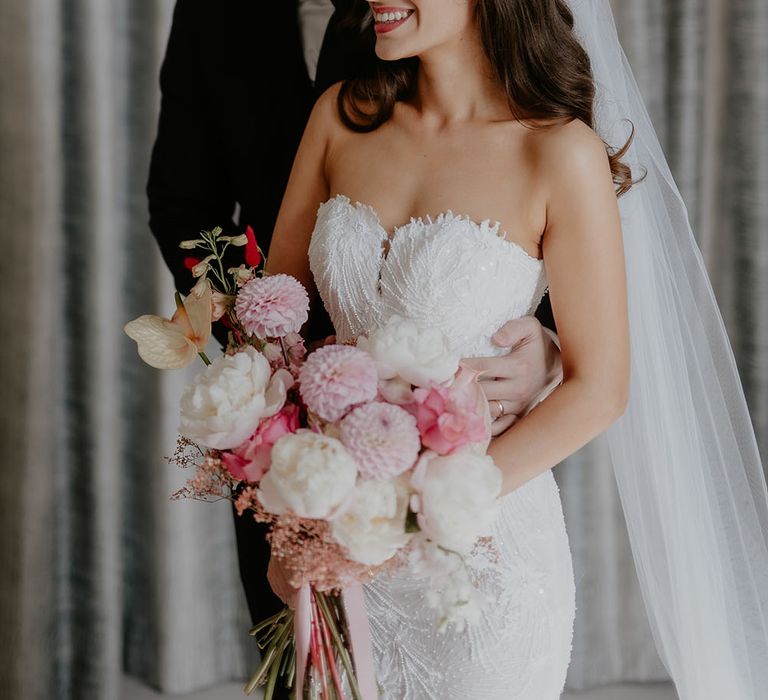 Groom stands behind his bride whilst she holds white and pink bridal bouquet and wears Pronovias wedding dress