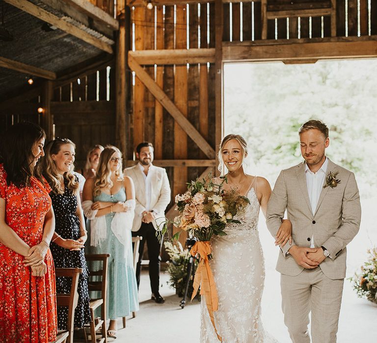 Bride smiles in a Made With Love wedding dress carrying her bouquet tied with orange ribbon as she is walked down the aisle 