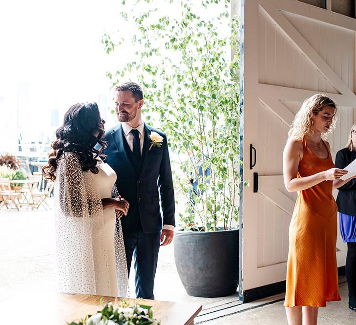 Bride & groom stand within Trinity Buoy Wharf as wedding guest gives reading on their wedding day