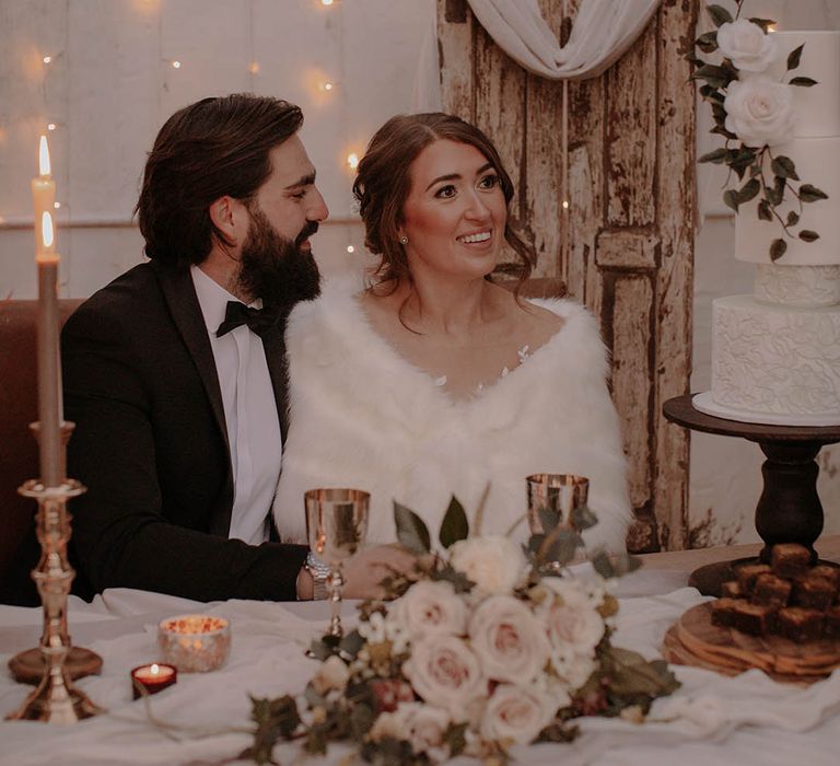 Bride wears a white fur shawl and the groom wears black tie as they sit down in the evening at Barlow Woodseats Hall