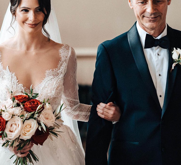 Father of the bride in a black tuxedo walks the bride in a long sleeve lace wedding dress down the aisle 