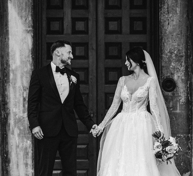 The groom in black tie stands smiling with the bride in a long sleeve lace tulle wedding dress 