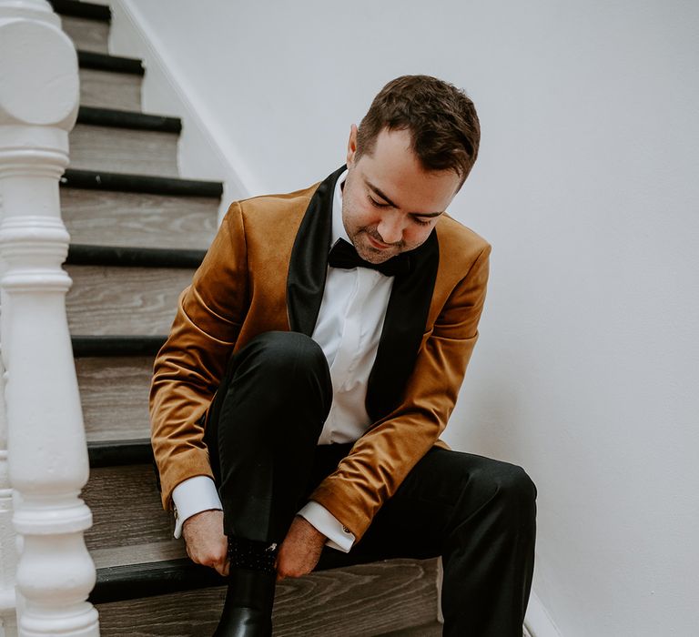 Groom in burnt orange velvet blazer puts on black boots while sitting on staircase