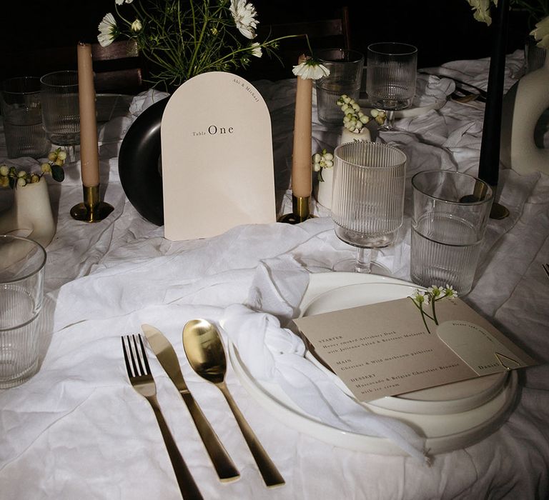 Monochrome minimalistic wedding tablescape with rounded off-white table number, gold cutlery, tan tapered candles and black and white table decorations