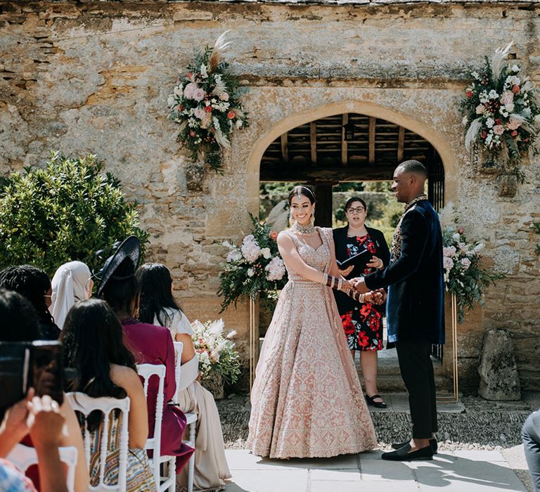 Panjabi bride and Jamaican Groom hold hands at alter wearing golden bridal Lehnga and midnight blue and gold Indian groom outfit