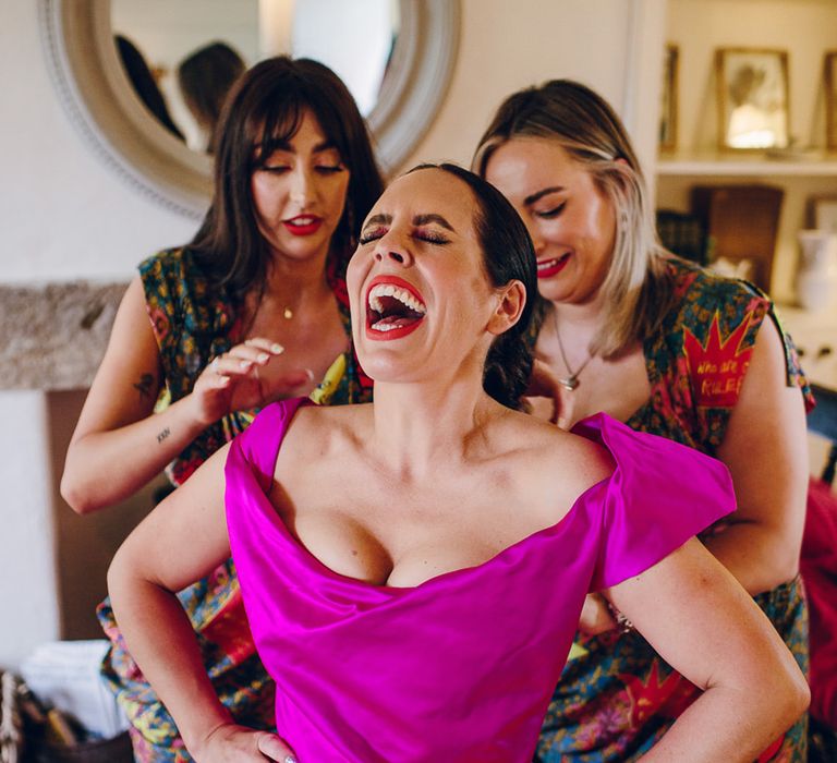 Bride in off the shoulder front ruching pink wedding dress and two bridesmaids in matching midi green colourful patterned Vivienne Westwood bridesmaid dresses helping her