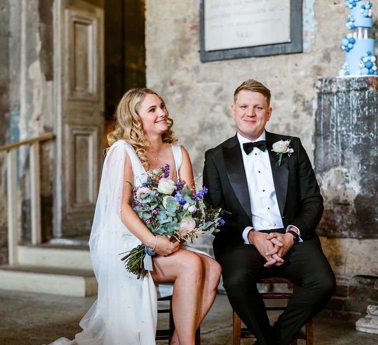 Bride holds colourful floral bouquet and sits beside her groom in black tie during wedding ceremony at The Asylum Chapel