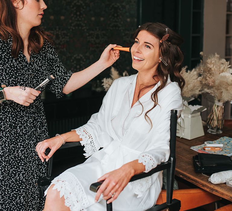 Bride in white robe with crochet trim sits getting her makeup done for her wedding day 