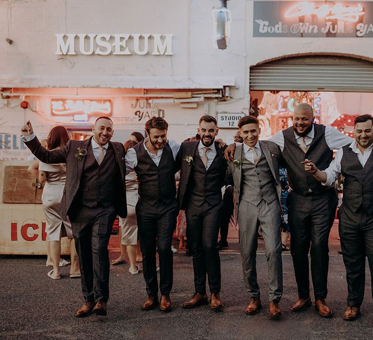 Groom in grey three piece wedding suit with yellow suit and yellow pocket square with groomsmen matching dark grey three piece suits with yellow ties and boutonnieres at Gods Own Junkyard 