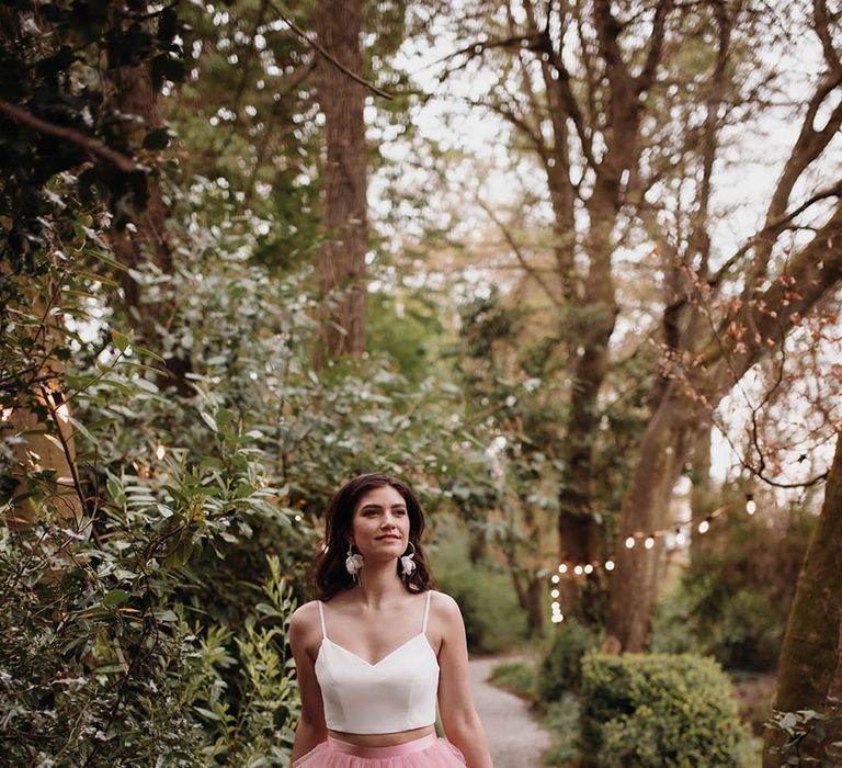 Bride wears white sweetheart neckline strappy crop top with ruffled pink skirt with high-low styling and silk waistband 