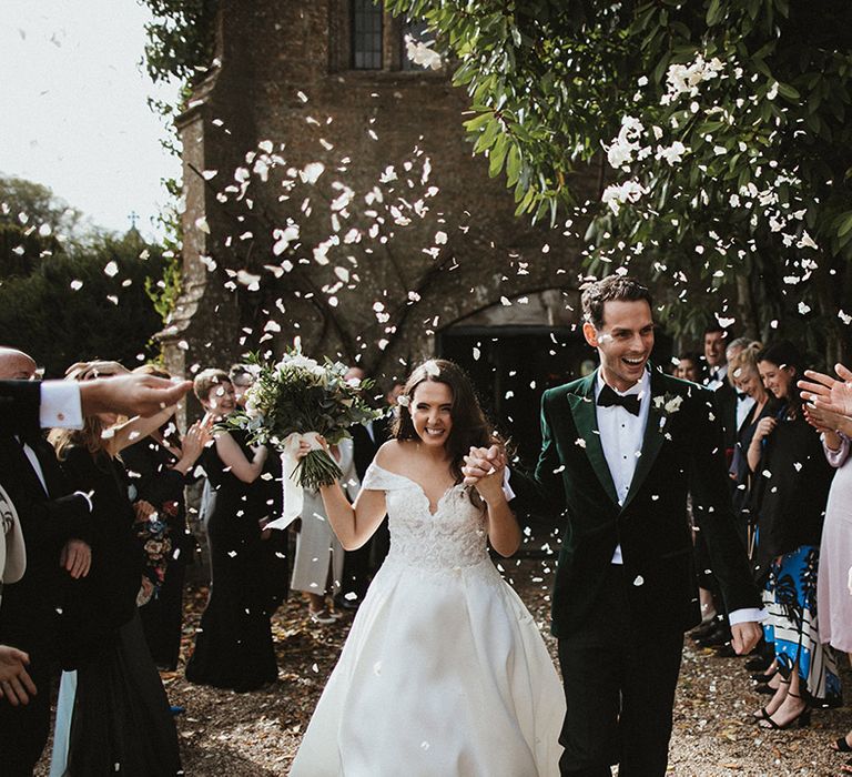 Bride in off the shoulder lace wedding dress walking with the groom in a green velvet tuxedo walk out of their ceremony to confetti 