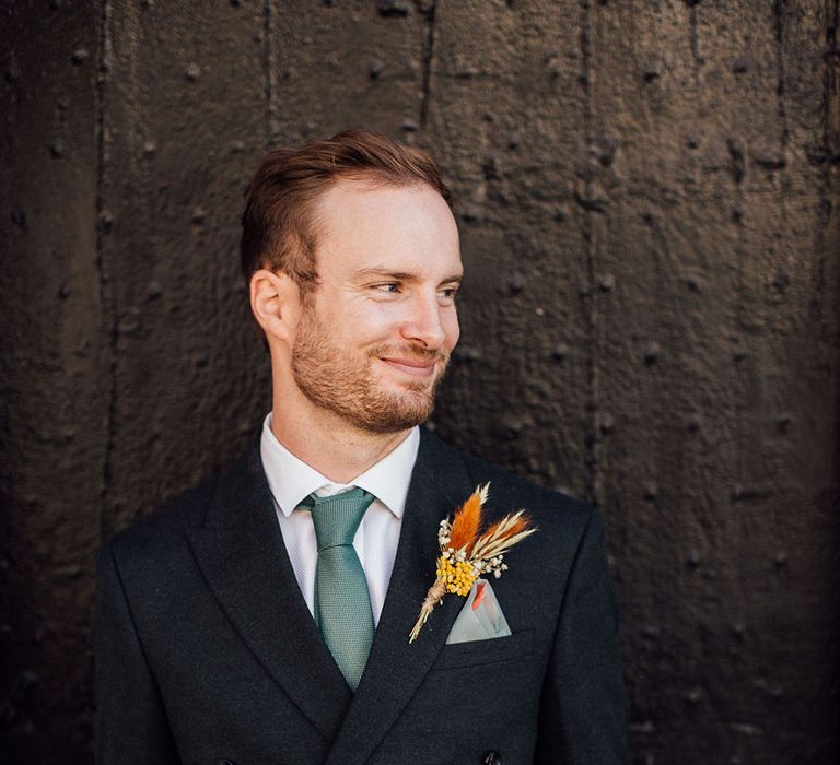 Groom wears suit complete with green tie and colourful pampas grass floral buttonhole 