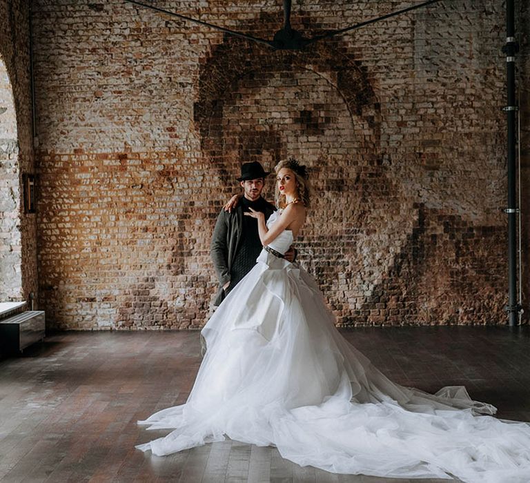 Bride in romantic organza and tulle a-line wedding day bridal ballgown with a flowing skirt, sculpted bandeau top and patterned black belt with dramatic, chunky brass necklace standing with groom in dark grey jacket, black turtle neck, black trousers and a black fedora hat 