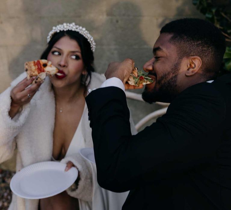 Bride in v neck wedding dress, white faux fur coat and large pearl headband eating pizza with groom in classic black tuxedo 