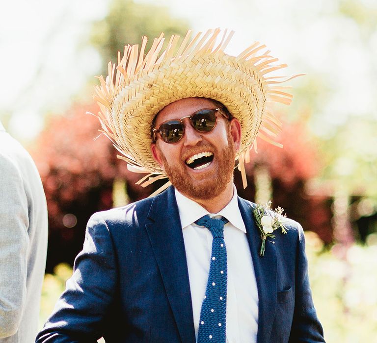 Groom in blue suit jacket with spotted tie wearing a big straw hat and sunglasses for outdoor summer wedding 