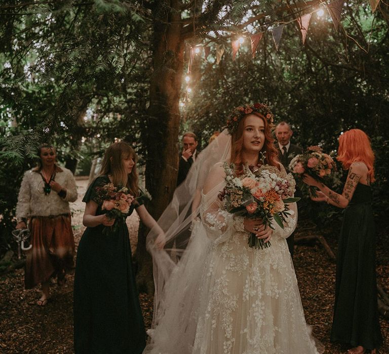 Bride in floral lace boho wedding dress gets ready to walk down the aisle with the bridesmaids in green bridesmaid dresses and the father of the bride 