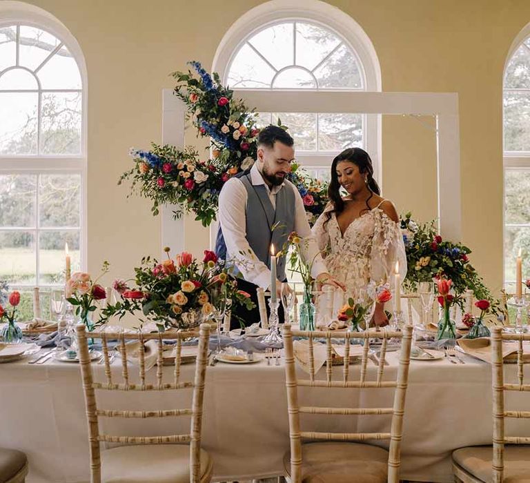 Bride in v-neck long sleeve shoulder cut out 3d appliqué wedding dress with flecks of gold standing with groom in cornflower blue waistcoat and black suit trousers by classic wedding tablescape with white wedding tablecloth, cornflower blue wedding table runner, bright blue glass bottles with dried flowers, pastel tapered candles and spring floral arrangements with suspended spring floral decor 