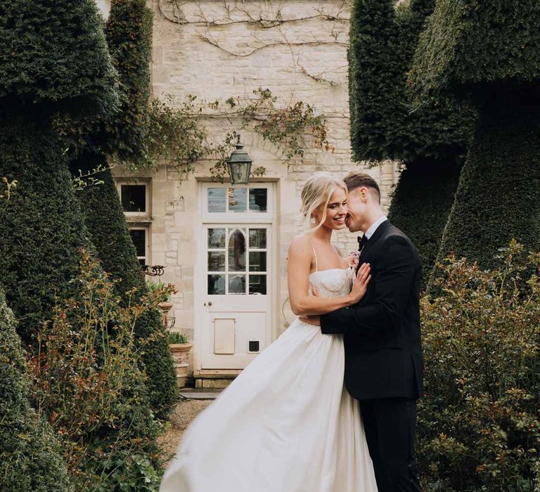 Bride in Watters strappy sleeveless princess wedding dress and chapel length wedding veil and groom in classic black tuxedo embracing on the grounds of Euridge Manor 