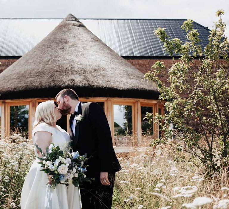 Bride in satin off the shoulder wedding dress holding white and blue eucalyptus, foliage, garden rose, gardenia, light blue delphinium and dried flower bridal bouquet tied with dusky blue ribbon kissing groom in classic black grooms three piece tuxedo with white rose boutonniere and black bowtie outside of Brickhouse Vineyard Devon wedding venue