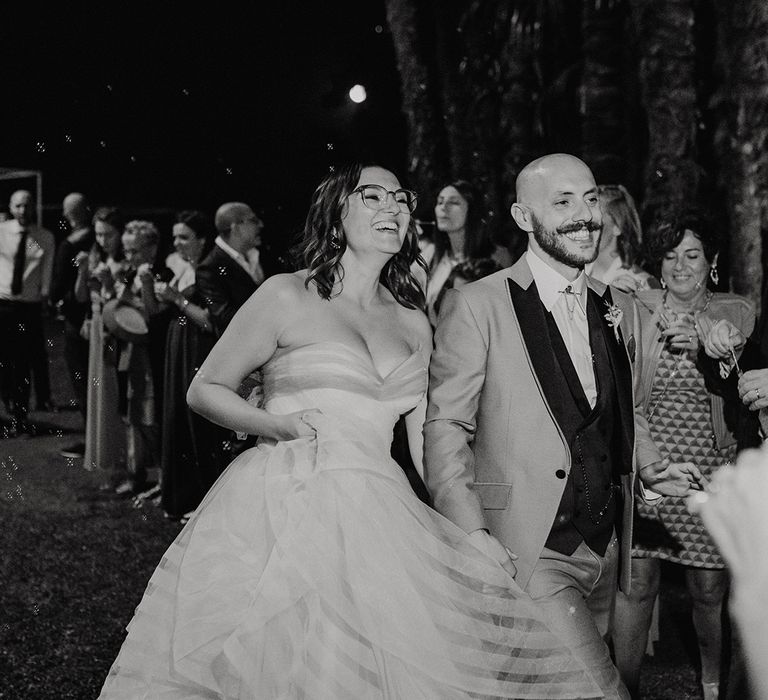 bubble wedding exit with bride in strapless wedding dress with layered skirt and groom in a light grey suit with dark lapel and waistcoat