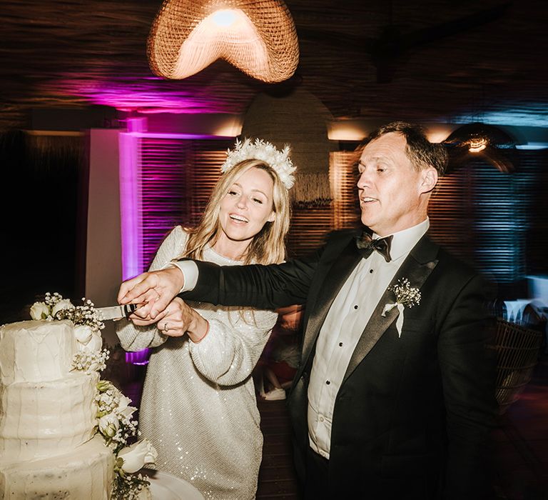 bride in a short sparkly wedding reception dress with long sleeves wearing a flower headband cutting the wedding cake with her groom in a tuxedo and bow tie