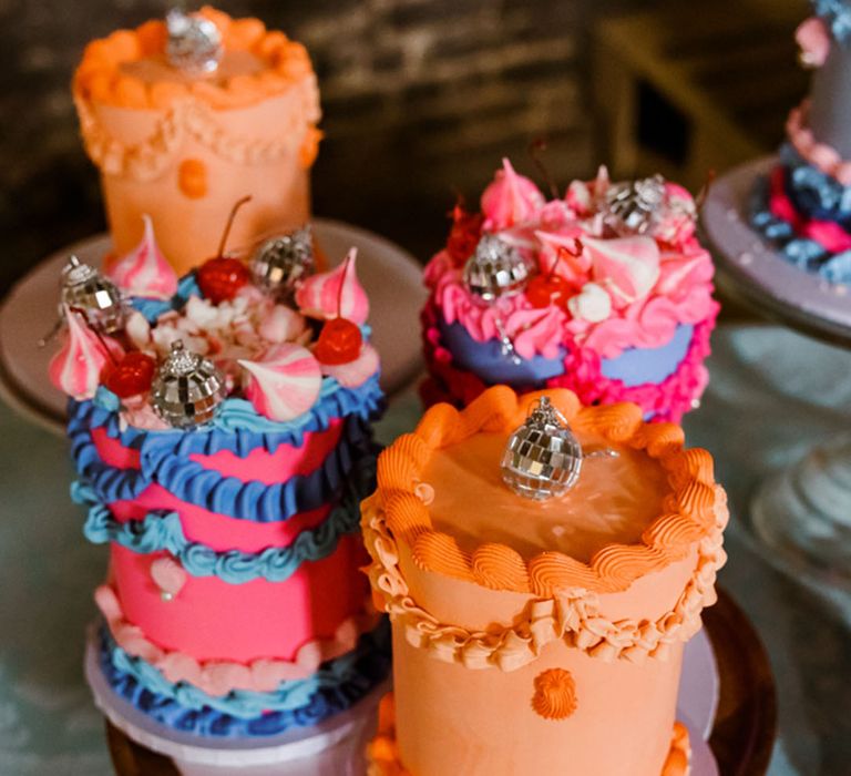 A table full of mini retro wedding cakes in bright shades of orange, pink and blue decorated with mini disco baubles 