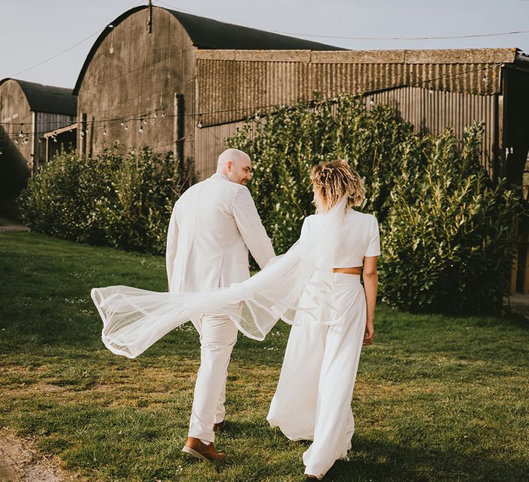 Bride in stunning bridal trouser two piece set with veil walking hand in hand with the groom 