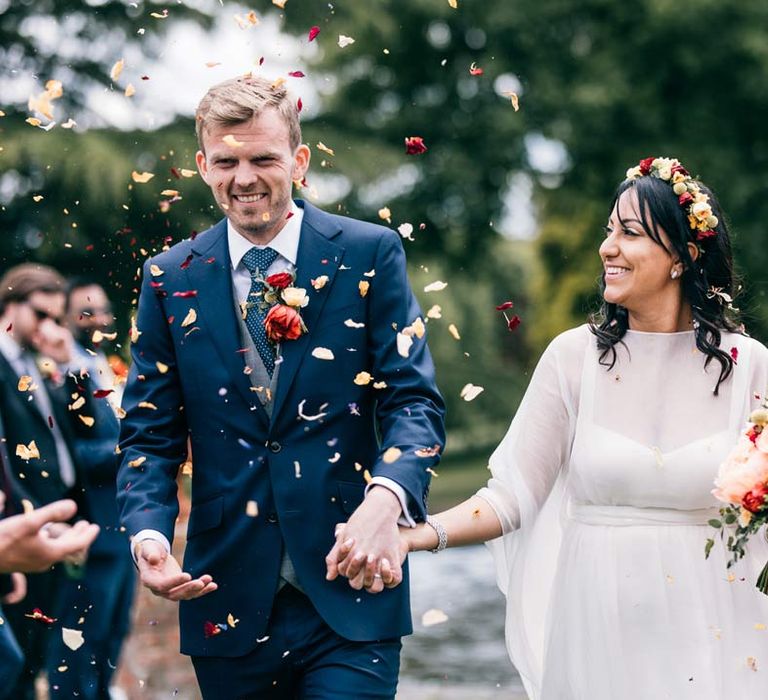 Groom in classic navy grooms suit with black tie, grey waistcoat and colourful rose boutonniere walking with bride in long sleeve mesh overlay wedding dress and colourful bridal flower crown doing colourful dried petal confetti exit 