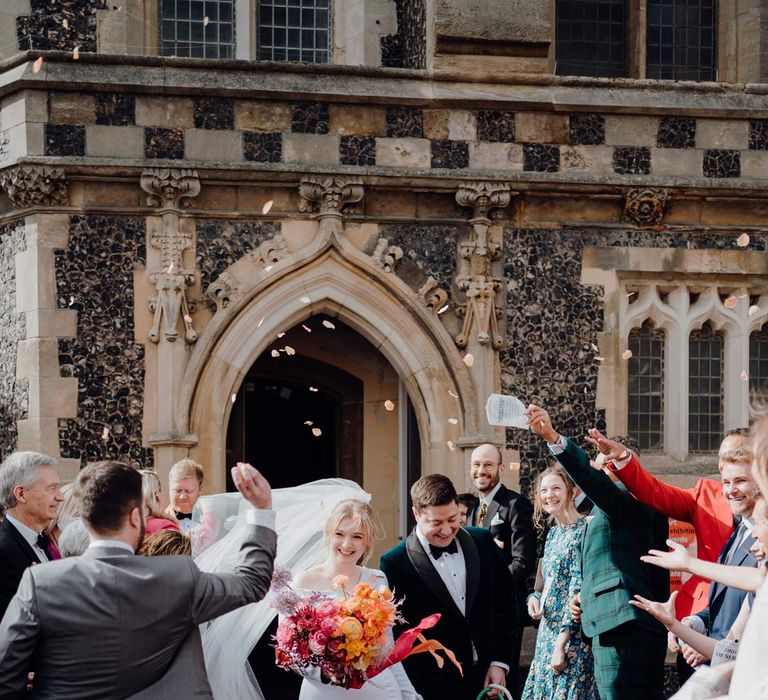 Bride in asymmetrical off shoulder long sleeve satin wedding dress with cathedral length personalised veil holding colourful bridal bouquet with pink garden roses, purple giant alliums, dark red poppies and foliage doing colourful confetti exit with groom in classic tuxedo and black bowtie with their puppy