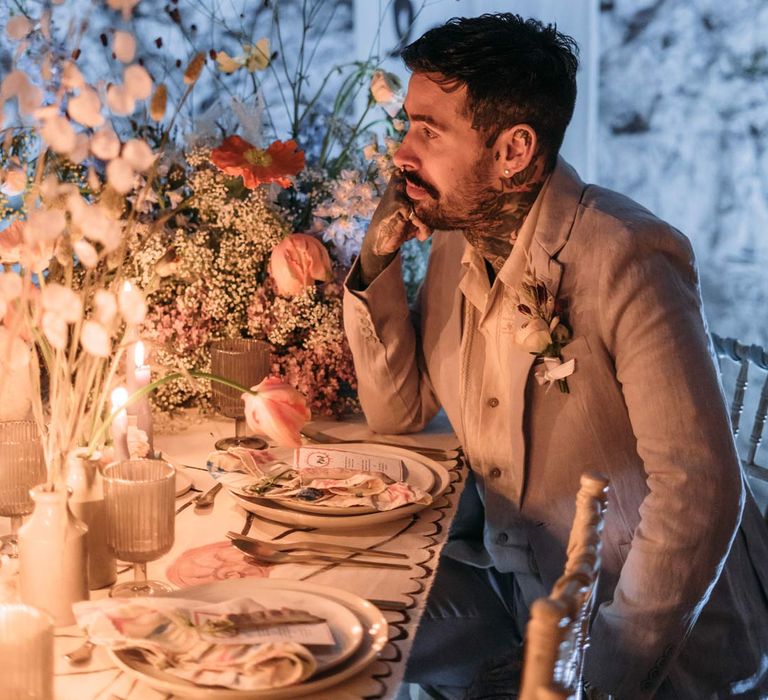 Groom in beige linen suit and embroidered linen shirt on the steps at Margate Caves sitting by artsy abstract wedding tablescape and alternative wedding decor with gold cutlery, off-white plates and abstract wedding stationery