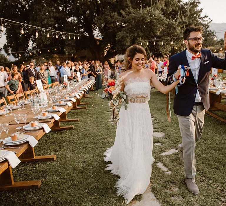 groom in grey chinos navy blazer and orange bow tie holding hands with his bride in a strapless lace wedding dress during their outdoor wedding reception