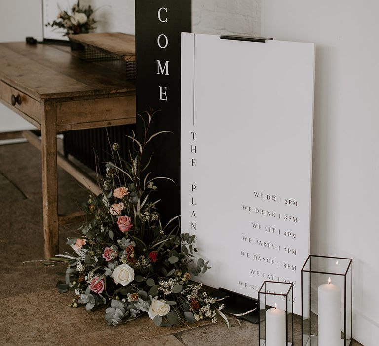 Tall vertical black and white wedding welcome sign with black and white order of the day sign surrounded by pink wedding flowers 