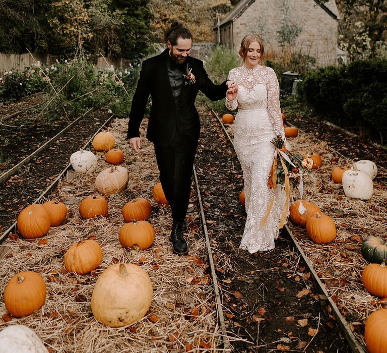 Bride in long sleeve boho lace wedding dress with groom walking through pumpkin patch 