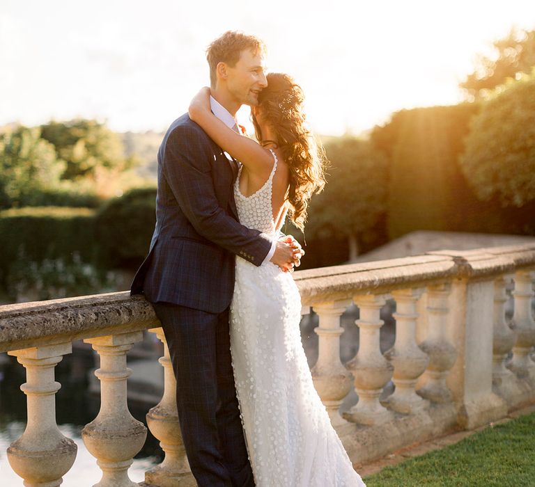 golden hour photograph with groom in a navy suit embracing his bride in a fitted appliqué wedding dress