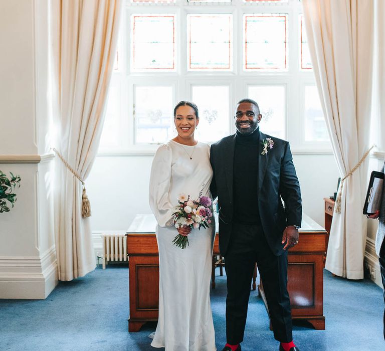 Groom in black suit with bright red socks standing eight the bride in a long sleeve satin wedding dress holding purple bouquet 