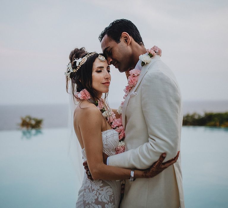 Boho bride in white lace wedding dress with flower hair accessories and traditional Matha Patti standing in front of swimmng pool with groom in groom suit and both wearing traditional pink Varmalas