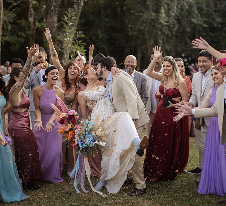 The wedding party wearing bright colourful and purple wedding outfits celebrate as the bride and groom kiss 