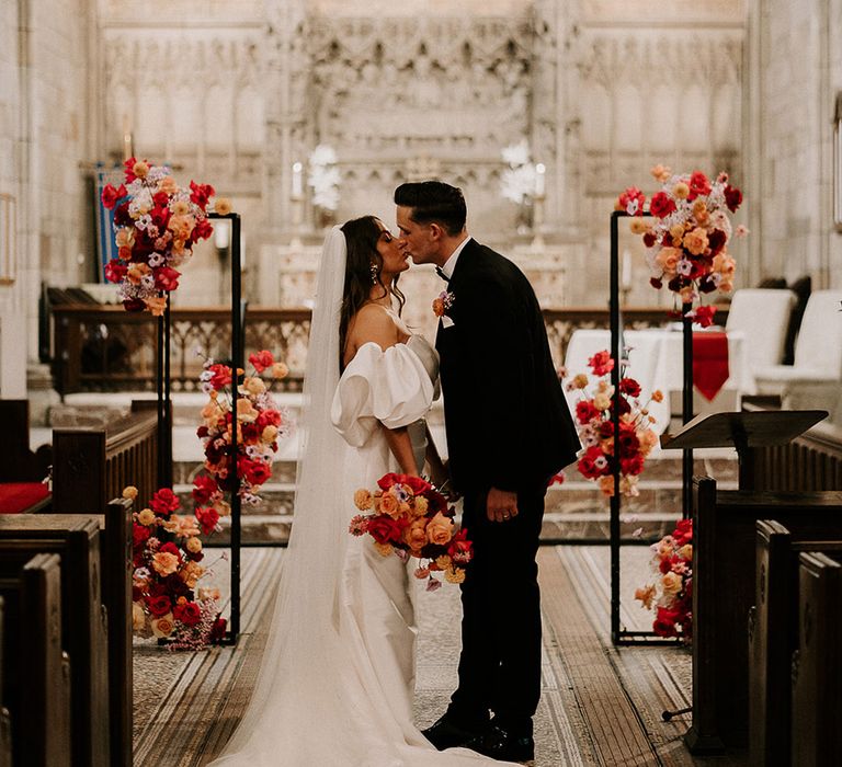 Bright and bold colourful wedding flowers decorate the church as the bride and groom kiss 