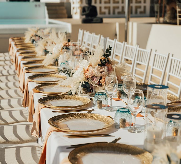 Destination wedding tablescape with gold and coral touches including gold charger plates and pampas grass and peach flower arrangements