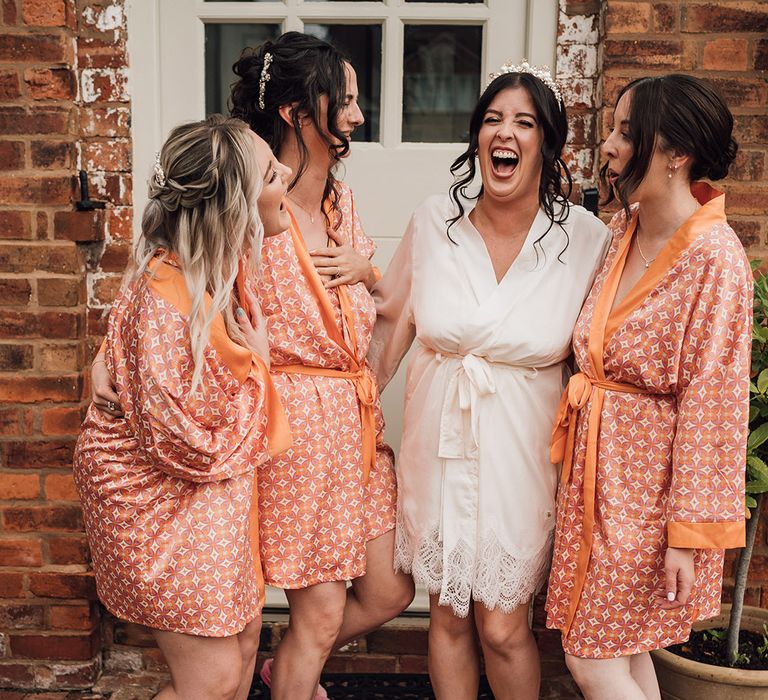 Bridesmaids wearing patterned orange satin robes with the bride in a white bridal robe as they get ready 