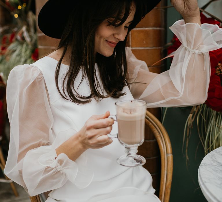 Bride in Knee length wedding dress and fedora hat enjoying a coffee at intimate wedding